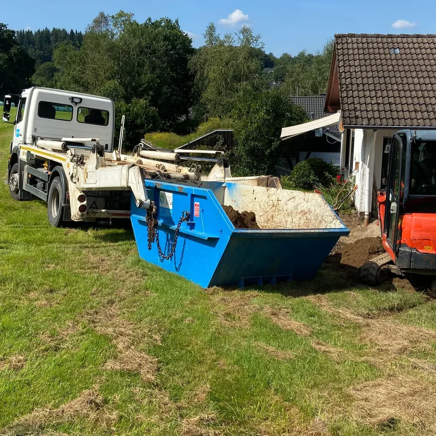Absetzkipper stellt einen blauen Container neben einem Wohnhaus ab, daneben der Minibagger